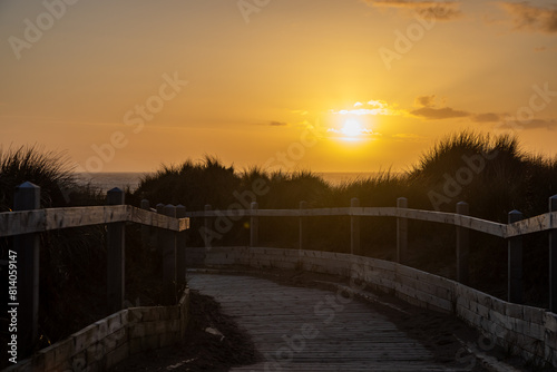 A wooden boardwalk leading through marram grass covered sand dunes to the beach  with a sunset sky overhead