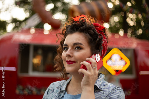 Closeup photo. Young adult beautiful pinup woman with curly hair is calling on mobile phone with interest. Cute girl at street food festival near foodtruck. Sunny day. Communication, negotiation, call