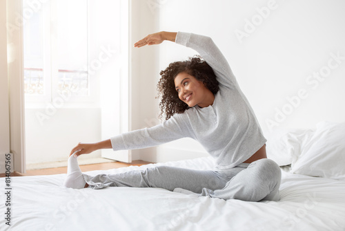Hispanic woman is stretching her body while lying on a bed with white sheets. She is extending her arms and legs to loosen up her muscles after waking up. photo