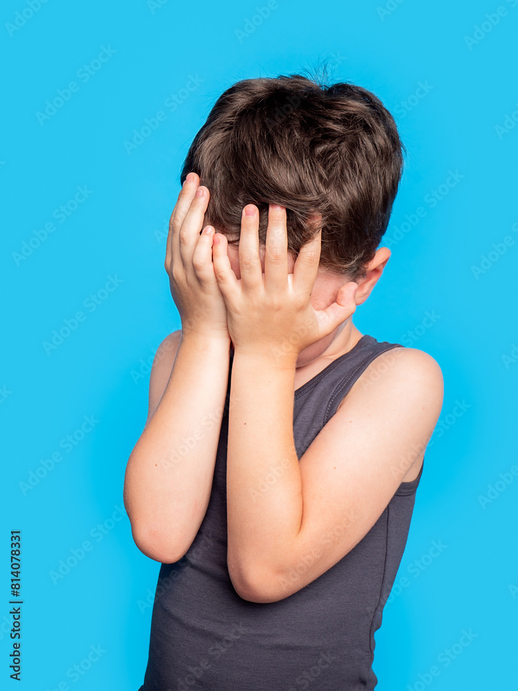 Young boy hiding face with hands on blue background Young boy hiding face with hands on blue background