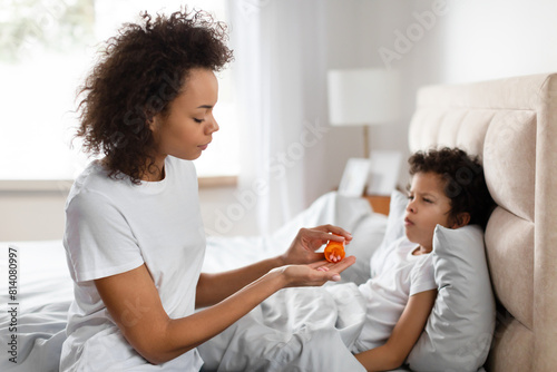 A concerned African American mother sits beside her bedridden young child, carefully administering oral medication. The natural light from a nearby window illuminates the serene bedroom