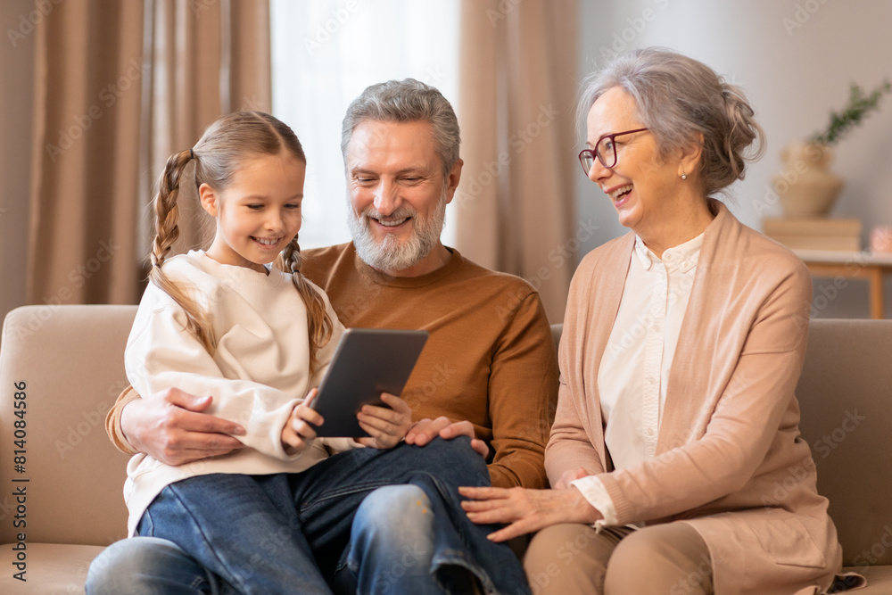A family consisting of parents and children sitting together on a couch, engaged with a tablet device. They are focused on the screen, interacting with the content displayed. A family consisting of parents and children sitting together on a couch, engaged with a tablet device. They are focused on the screen, interacting with the content displayed.