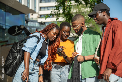 Interested stylish group of african american young people students looking at smartphone while walking in street. Happy black woman showing something to joyful friends in cellphone have fun outdoors.
