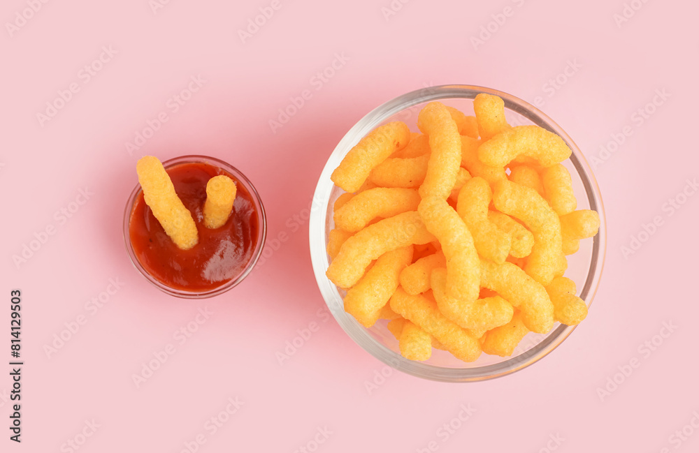 Glass bowl with tasty salty corn sticks and ketchup on pink background