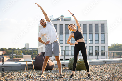 Happy sports couple making health gymnastics in morning on roof terrace of building. Positive man in white t-shirt and woman in black top doing sports stretching their arms up.
