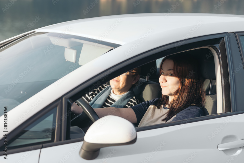 Naklejka premium A young girl learns to drive a car with her mother in the passenger seat as the sunlight illuminates their smiling faces.