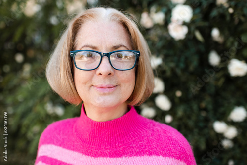 Smiling blonde mature woman 50-60 year old with gray hair wearing vision glasses and pink knitted sweater over blooming rose flowers outdoor. Looking at camera.