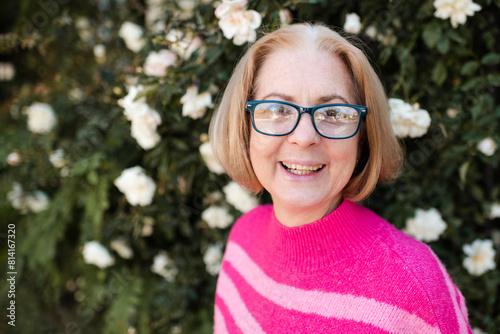 Smiling blonde mature woman 50-60 year old with gray hair wearing vision glasses and pink knitted sweater over blooming rose flowers outdoor. Looking at camera.