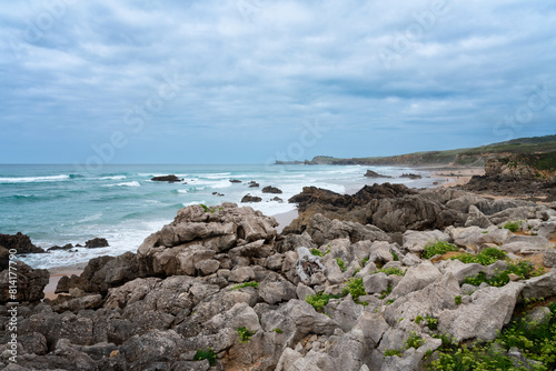 A rocky shoreline with a large body of water in the background. The rocks are scattered throughout the area. The sky is cloudy, giving the scene a dramatic mood. Liencres, Cantabria, Spain