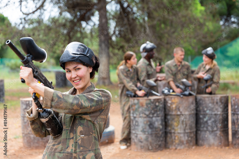 Fototapeta premium Smiling asian woman paintball player in camouflage posing with gun outdoors