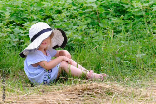 child 3 years old, girl in white hat, dress sit on grass in field, smiling trustingly, concept of young entomologist, naturalist, interest of preschoolers in study of plants, unity with nature photo