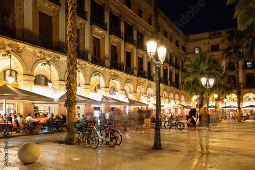 Evening view of bustling Placa Reial in Barcelona with well-lit cafes and people enjoying leisurely stroll. Favorite place for locals and tourists alike