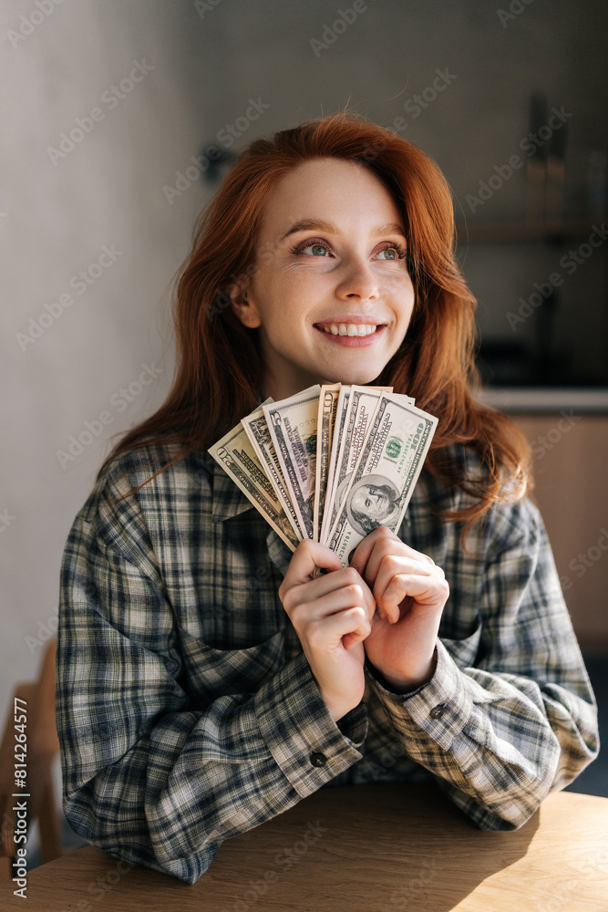Happy young woman counting money in dollars banknotes sitting in kitchen table at home. Rich happy female holding in hands stack of cash money of success, gain, victory. Concept of investment. Happy young woman counting money in dollars banknotes sitting in kitchen table at home. Rich happy female holding in hands stack of cash money of success, gain, victory. Concept of investment.