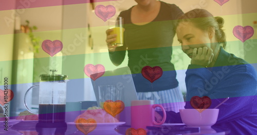 Image of heart emojis and rainbow flag over caucasian female couple at home