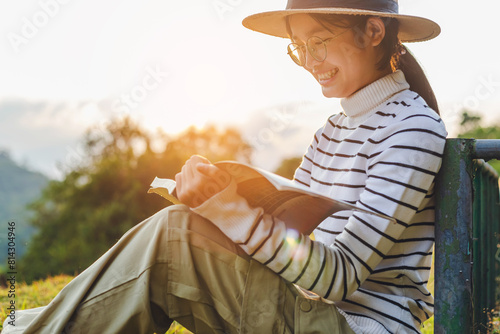 Side view cute girl with eyeglasses and hat sitting on grass and reading book in park sunset