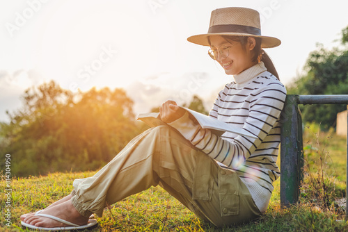 Side view cute girl with eyeglasses and hat sitting on grass and reading book in park sunset
