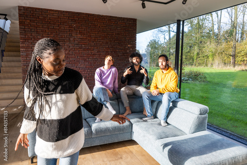 This image depicts a diverse group of four friends in a modern living room. A Black woman, standing, is interacting with three seated men, one Black, one White, and one Middle Eastern. The setting