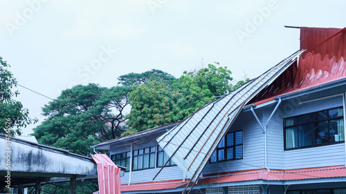 Broken roof after a storm. The roof of a wooden building covered with metal sheets is broken and damaged by a summer storm in Thailand on the background of green trees and hazy sky. Selective focus