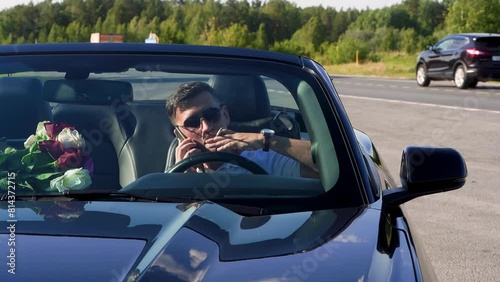Smiling man talking on smartphone while smoking cigarette. Young male is relaxing in convertible car on sunny day. Motor vehicles are passing on road in background.