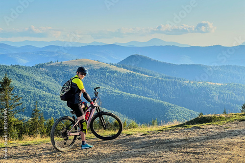 Cyclist man riding electric bike outdoors on sunny day. Male tourist resting on hill  enjoying beautiful mountain landscape  wearing helmet and backpack. Concept of active leisure.