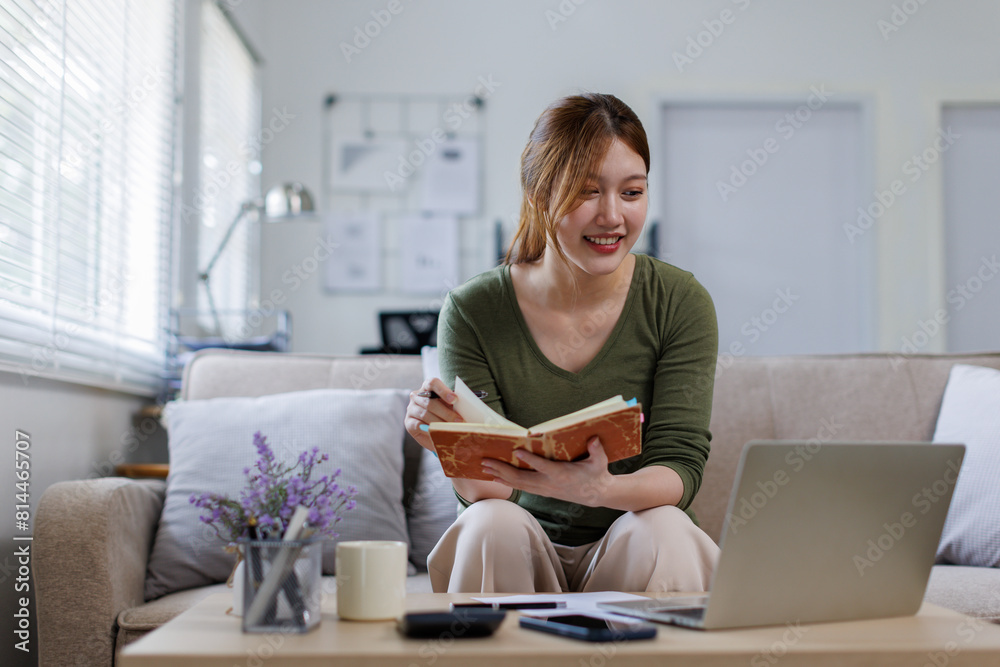 Young asian woman at home sitting on modern chair in front of window relaxing in her living room reading book and drinking coffee or tea Young asian woman at home sitting on modern chair in front of window relaxing in her living room reading book and drinking coffee or tea