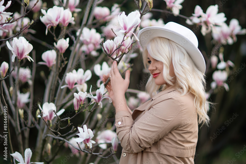 Fototapeta premium Magnolia flowers woman. A blonde woman wearing a white hat stands in front of a tree with pink flowers. She has a smile on her face and she is enjoying the beautiful scenery.