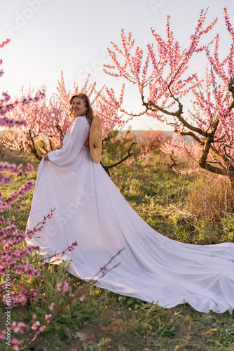 Woman blooming peach orchard. Against the backdrop of a picturesque peach orchard  a woman in a long white dress and hat enjoys a peaceful walk in the park  surrounded by the beauty of nature.
