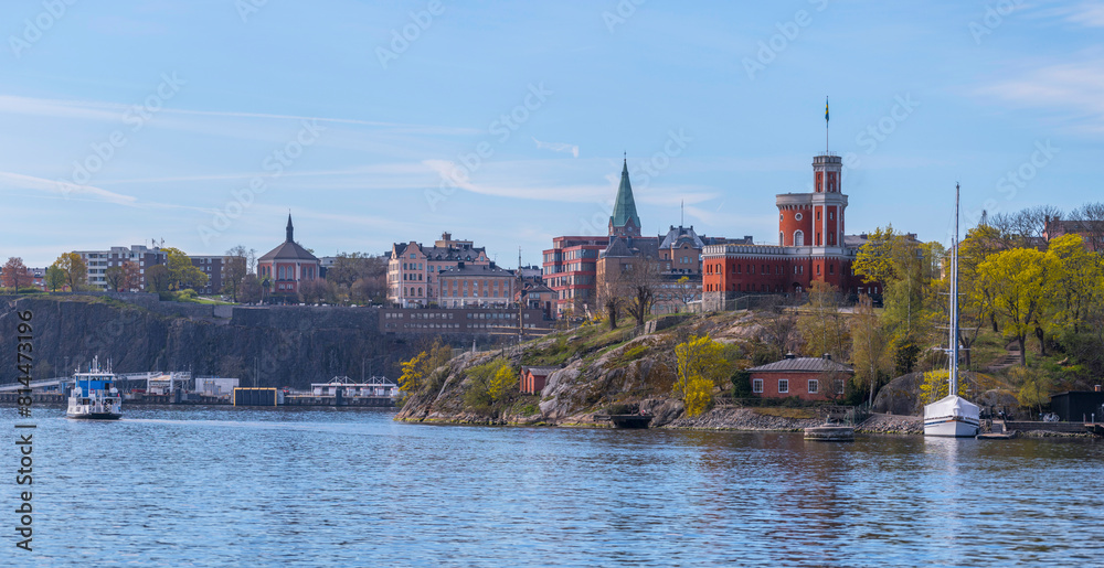 Fototapeta premium Small harbor ferry Djurgården passing the castle on the island Kastellholmen, the district Södermalm in the background, a sunny summer morning in Stockholm