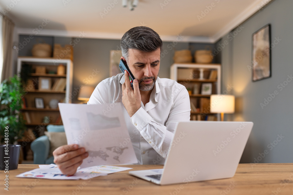 Handsome businessman working on laptop and talking on cellphone. Handsome mature business leader sitting in a modern office Handsome businessman working on laptop and talking on cellphone. Handsome mature business leader sitting in a modern office