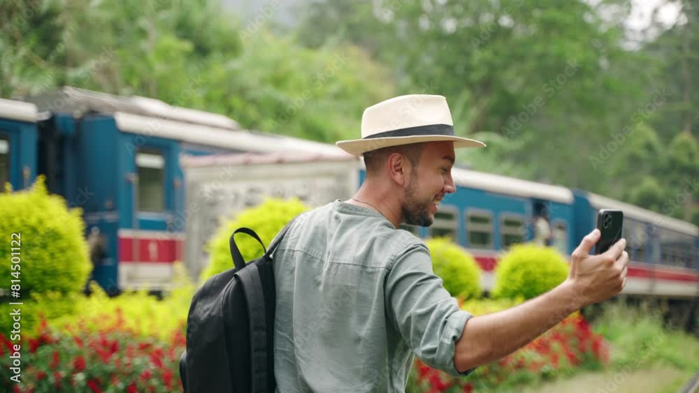 Asian travel blogger in hat records video with smartphone at tropical train station. Excited male tourist captures moving train, blooming flowers, surrounding greenery on camera, creates content. Asian travel blogger in hat records video with smartphone at tropical train station. Excited male tourist captures moving train, blooming flowers, surrounding greenery on camera, creates content.