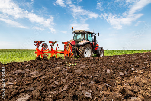 Red tractor plows through fertile soil with a vibrant green landscape in the background photo