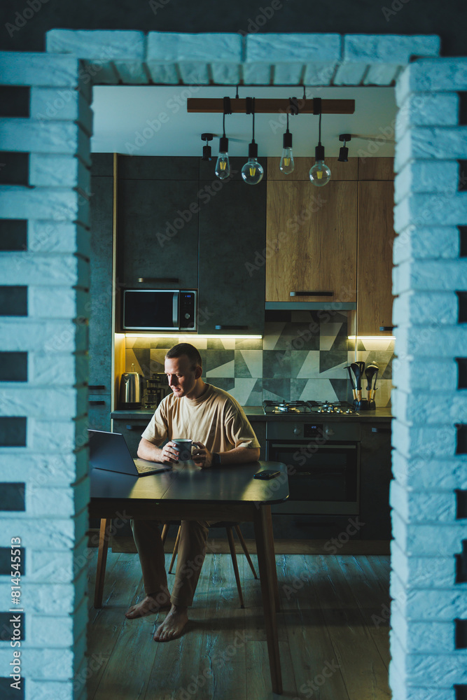 A young man sits at the table in the kitchen and works on a laptop. The laptop is on the table and the young man is working at home. A young man sits at the table in the kitchen and works on a laptop. The laptop is on the table and the young man is working at home.