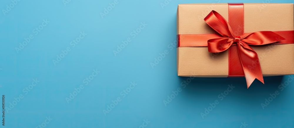A brown paper gift box with a red satin ribbon bow is placed at the bottom of a blue background The image is taken from a top down perspective offering copy space This composition embodies the concept A brown paper gift box with a red satin ribbon bow is placed at the bottom of a blue background The image is taken from a top down perspective offering copy space This composition embodies the concept
