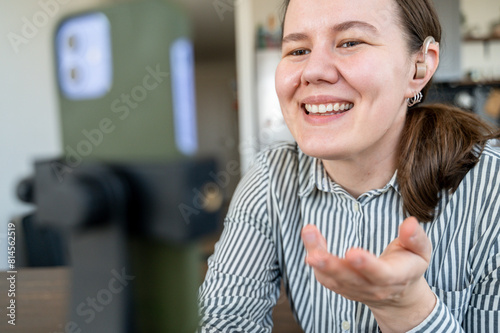 Portrait of young smiling woman with hearing aid on left ear recording video or making video call at home
