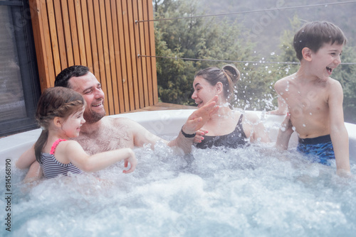 Happy family with two small children have fun in the jacuzzi. Cheerful young dad splashes water on his laughing cute son.