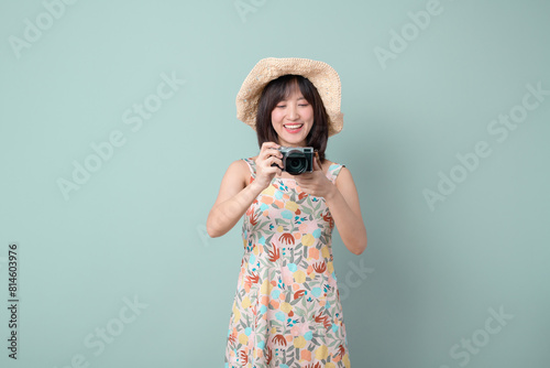 Happy Asian woman wearing casual dress and hat with camera on vacation or travel theme isolated on pastel green background. Travel and vacation concept.