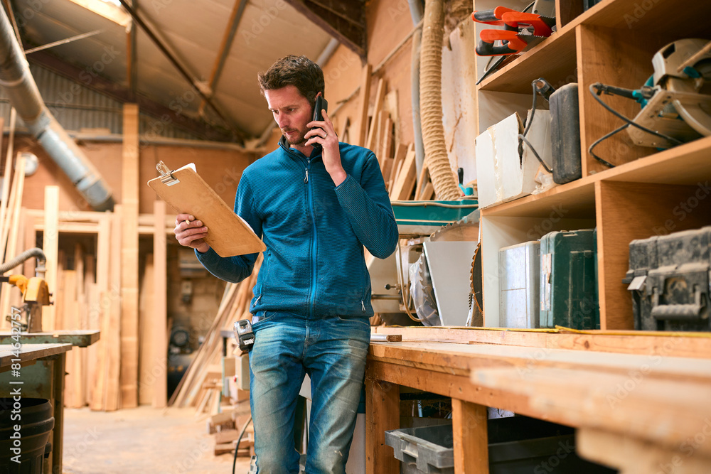 Carpenter Working In Woodwork Workshop Making Call On Mobile Phone Whilst Looking At Plan Carpenter Working In Woodwork Workshop Making Call On Mobile Phone Whilst Looking At Plan