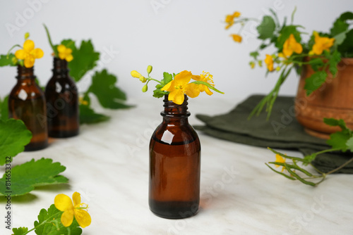 Celandine essential oil container with celandine flower and leaves on a white marble background. Selective focus. Side view. 