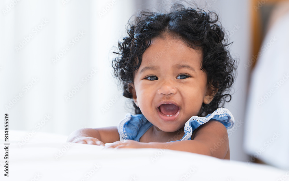 Portrait of adorable African 7 months old newborn baby girl with black curly hair standing near bed at home, looking at camera, smiling, laughing Portrait of adorable African 7 months old newborn baby girl with black curly hair standing near bed at home, looking at camera, smiling, laughing
