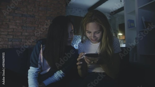 Happy friends sitting on sofa and looking at screen of mobile phone or smartphone. Quarantine using modern technology. Connecting through social networks or messages during epidemic. phubbing concept. photo