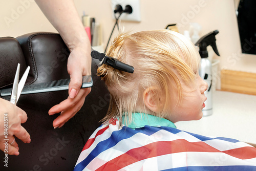 Little blond child boy getting haircut in beauty salon,barbershop. Hairdresser stylist cutting hair with professional tools-comb and scissors.Kids spa.Style and fashion,cutting hair for children
