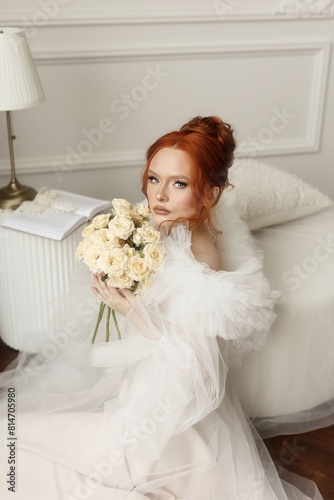 Radiant young woman with fiery red hair, dressed in white, striking a pose with a bouquet of white roses in a well-lit room. A red-haired young woman posing with a book and flowers.
