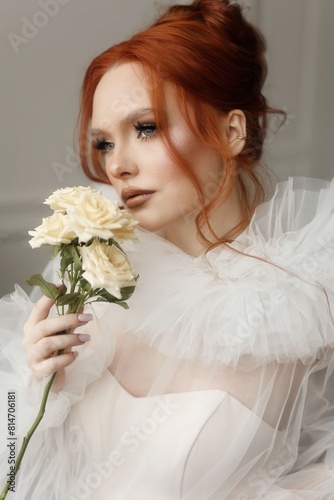 Graceful young woman with fiery red hair, dressed in white, posing with a bunch of white roses in a luminous interior. Portrait of a young woman in white.