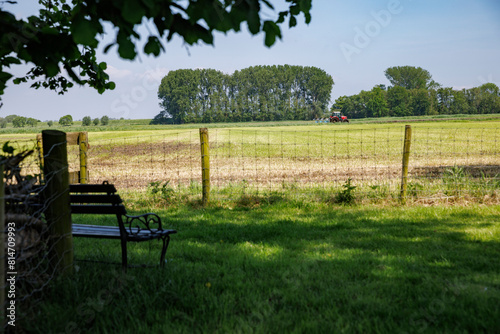 Tractor in a sunlit Dutch polder, with a shaded bench under trees.