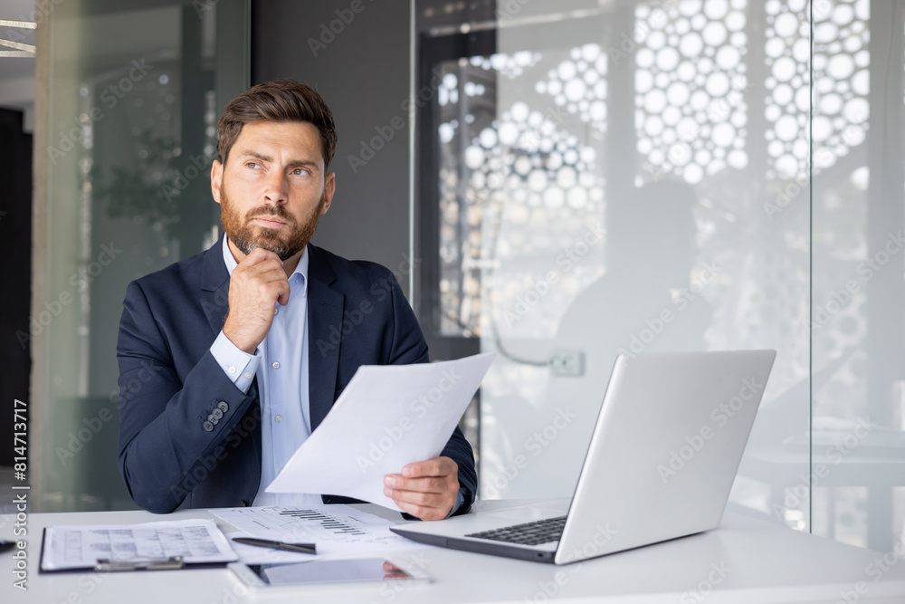 A serious and focused young man businessman works in the office, sits at a desk with a laptop, holds documents in his hands and looks thoughtfully to the side A serious and focused young man businessman works in the office, sits at a desk with a laptop, holds documents in his hands and looks thoughtfully to the side
