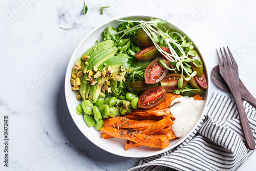 Vegan salad bowl with baked sweet potato, edamame beans, tomatoes, avocado, nuts, tahini dressing and sprouts.
