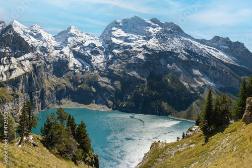 Oeschinensee lake with snow Bluemlisalp mountain on sunny summer day. Panorama of the azure lake Oeschinensee, pine forest in Swiss alps, Kandersteg. Switzerland.