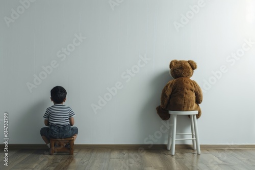 A poignant image of a young boy and his teddy bear sitting on stools facing a wall, illustrating a timeout scene. photo