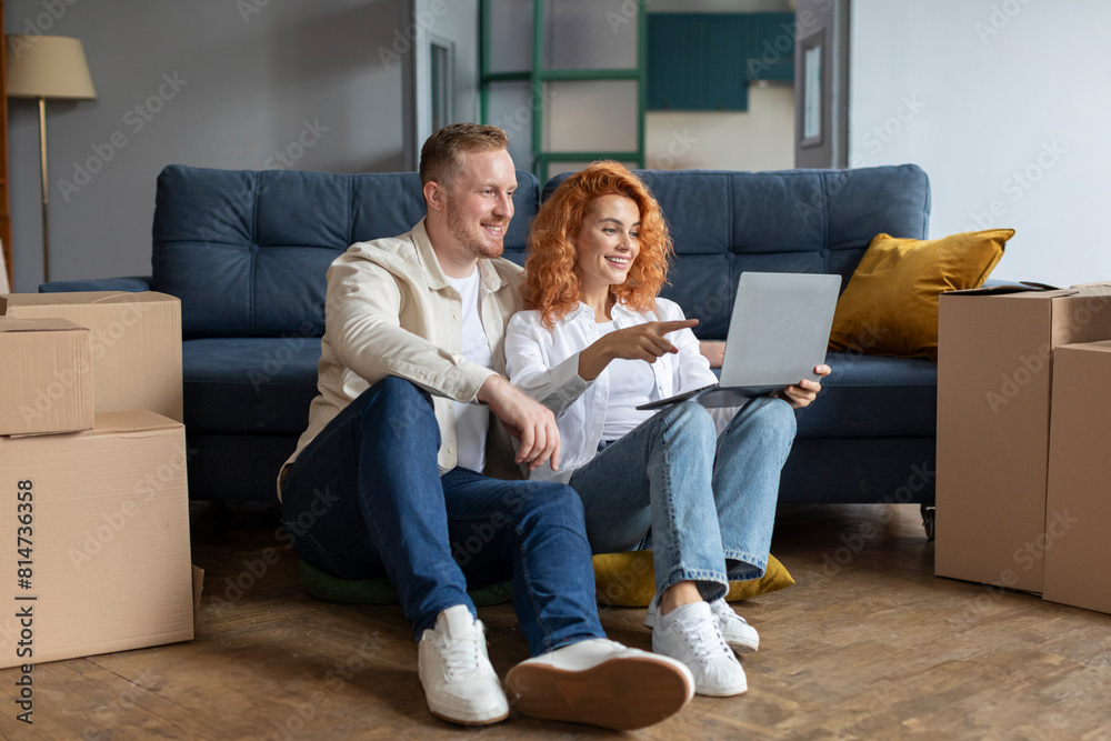 Couple using laptop computer sitting on floor among boxes after moving new home. Real estate, house ownership and relocation concept Couple using laptop computer sitting on floor among boxes after moving new home. Real estate, house ownership and relocation concept