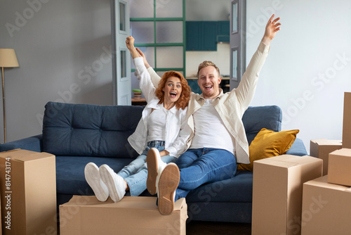 Excited husband and wife raise their hands up celebrating moving day, sitting on couch at new house. Rent real estate, buying home at mortgage © Home-stock